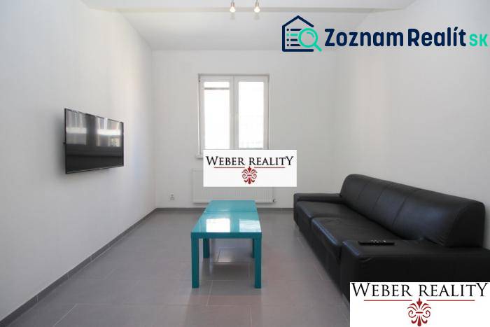 Wohnzimmer einer Zweizimmerwohnung mit schwarzem Sofa, Fernseher an der Wand und niedrigem Tisch.