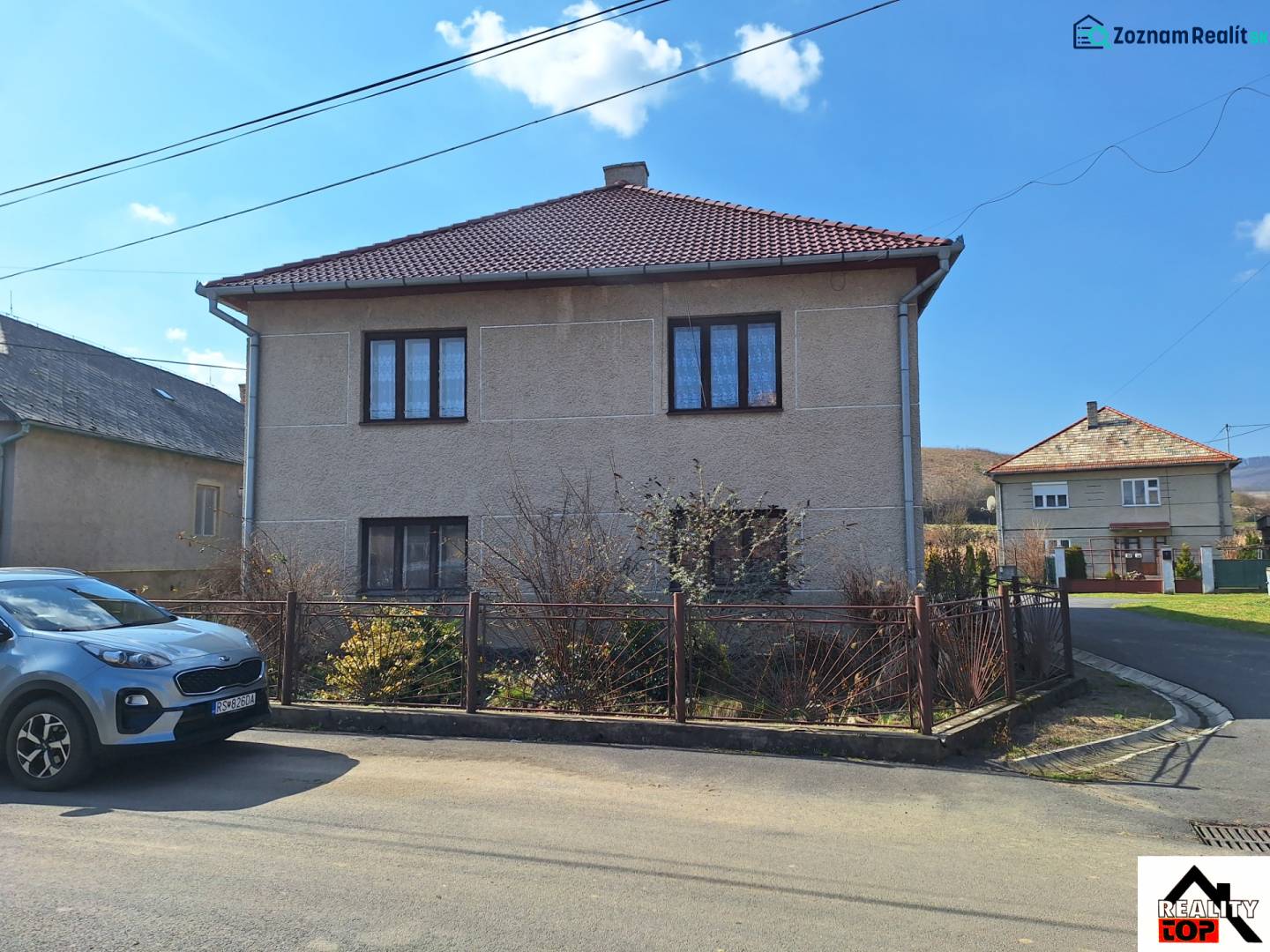 Ein Einfamilienhaus in Tachtoch mit einem Zaun, einem Auto und einem Nachbarhaus unter blauem Himmel.