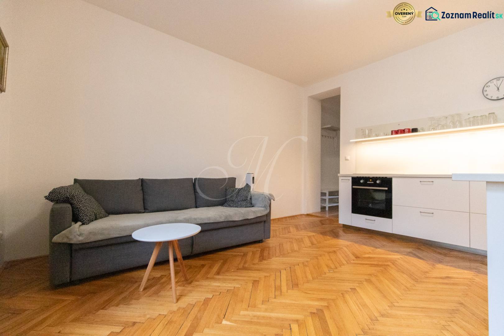 Wohnzimmer in einer Zweizimmerwohnung mit Sofa, Küchenzeile und Holzboden.