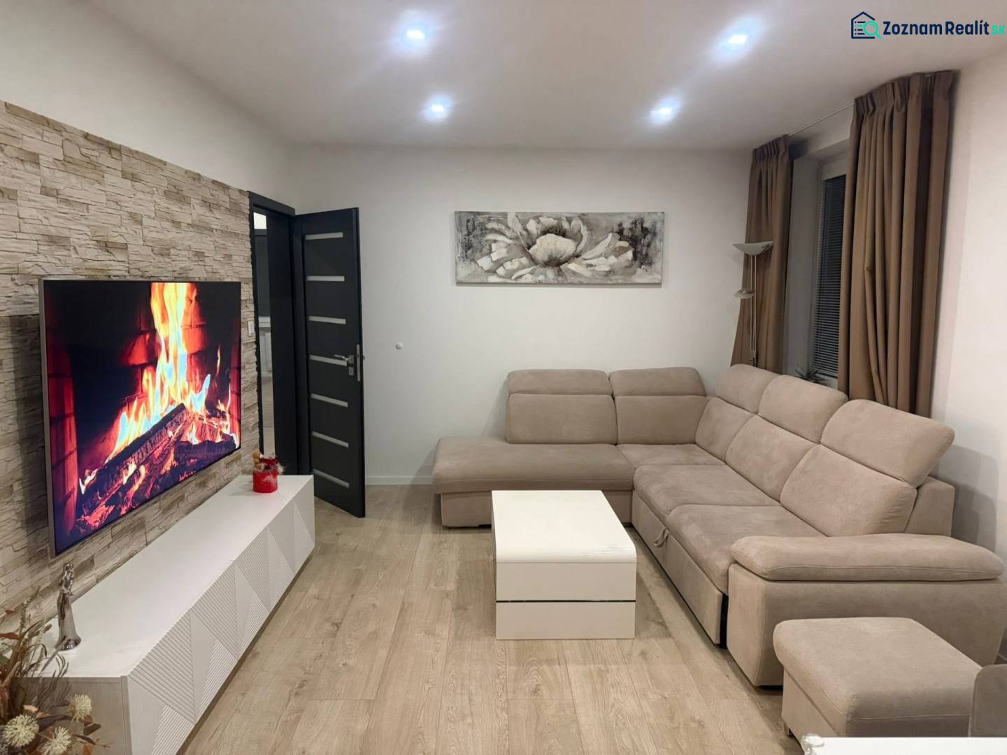 Wohnzimmer einer 3-Zimmer-Wohnung mit beigem Sofa, Fernseher und Boden in Holzoptik.