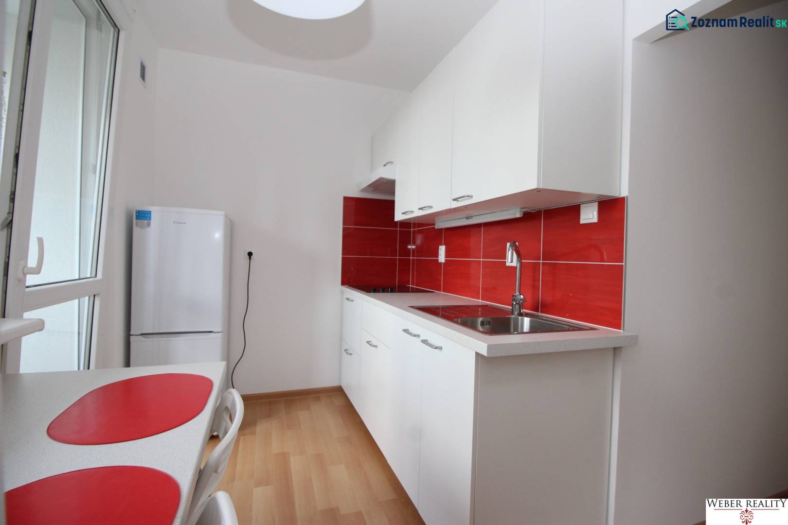Weiße Küche mit roter Fliesenverkleidung, Esstisch und Boden mit Holzdekor in einer Einzimmerwohnung.