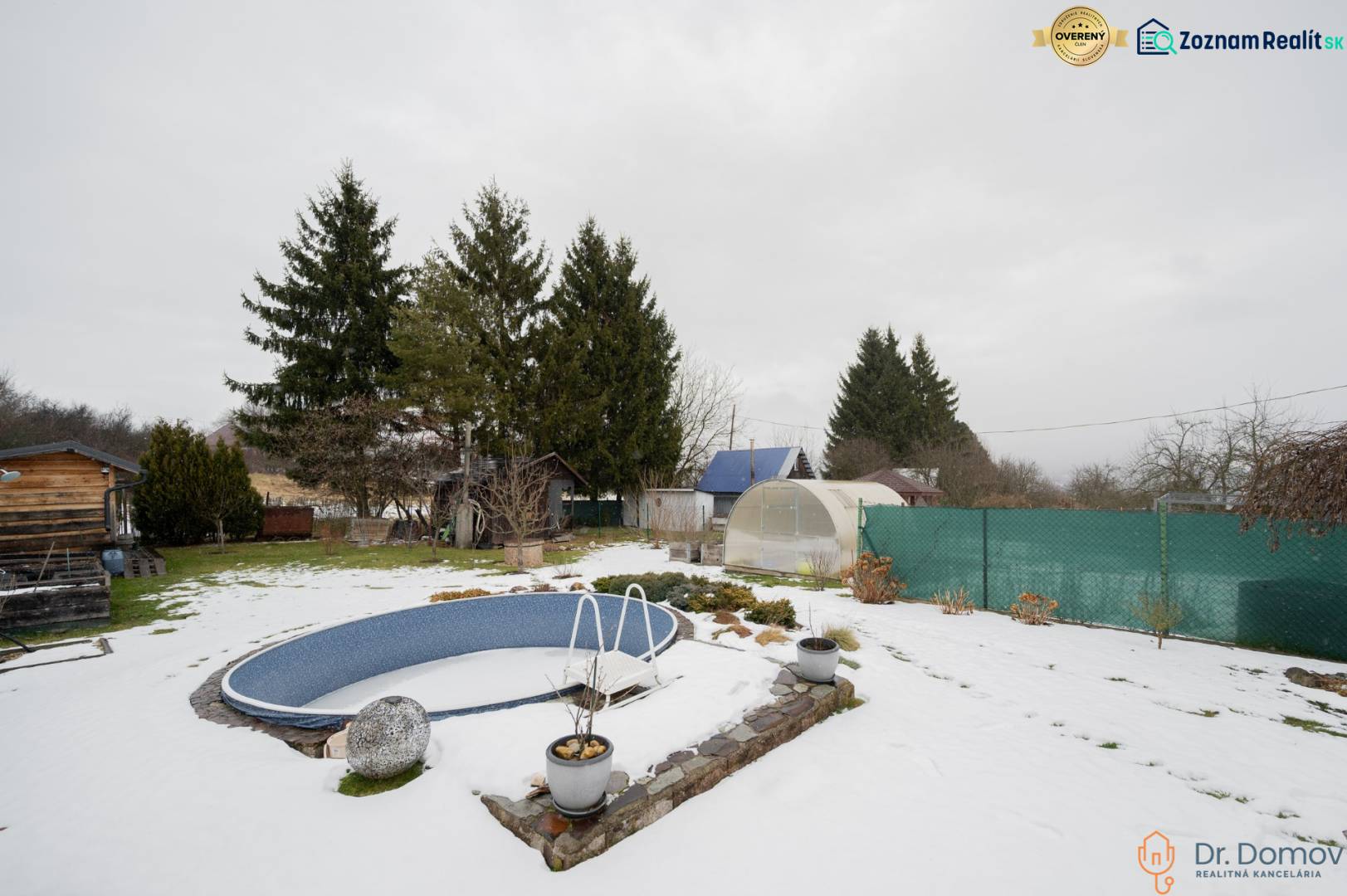 Ein verschneiter Garten bei einer Hütte in Handlová mit einem Pool, Gewächshäusern und Nadelbäumen.