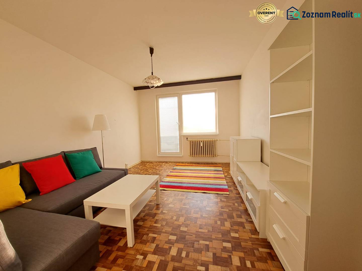 Wohnzimmer in einer 2-Zimmer-Wohnung mit einem Sofa, weißen Möbeln, bunten Kissen und einem Boden in Holzoptik.