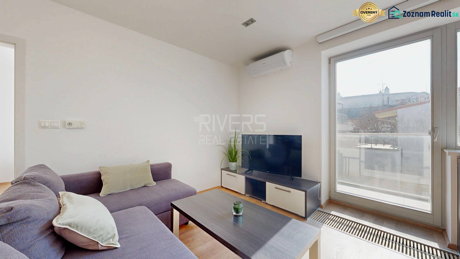 Wohnzimmer in einer Zweizimmerwohnung mit Sofa, Fernseher und großem Fenster.