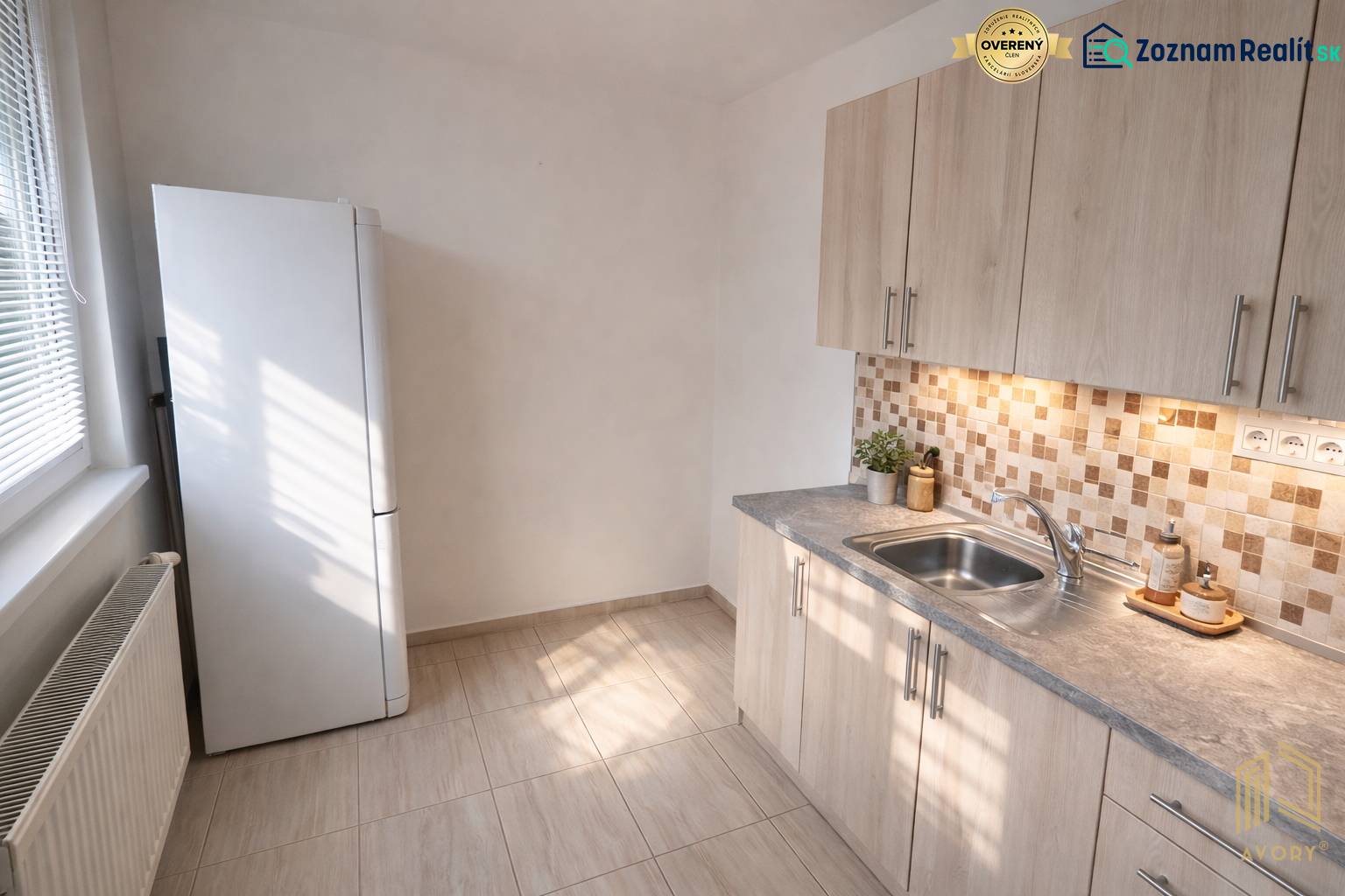 Küche in einer 2-Zimmer-Wohnung mit Fliesen und hellen Möbeln, einschließlich Kühlschrank.