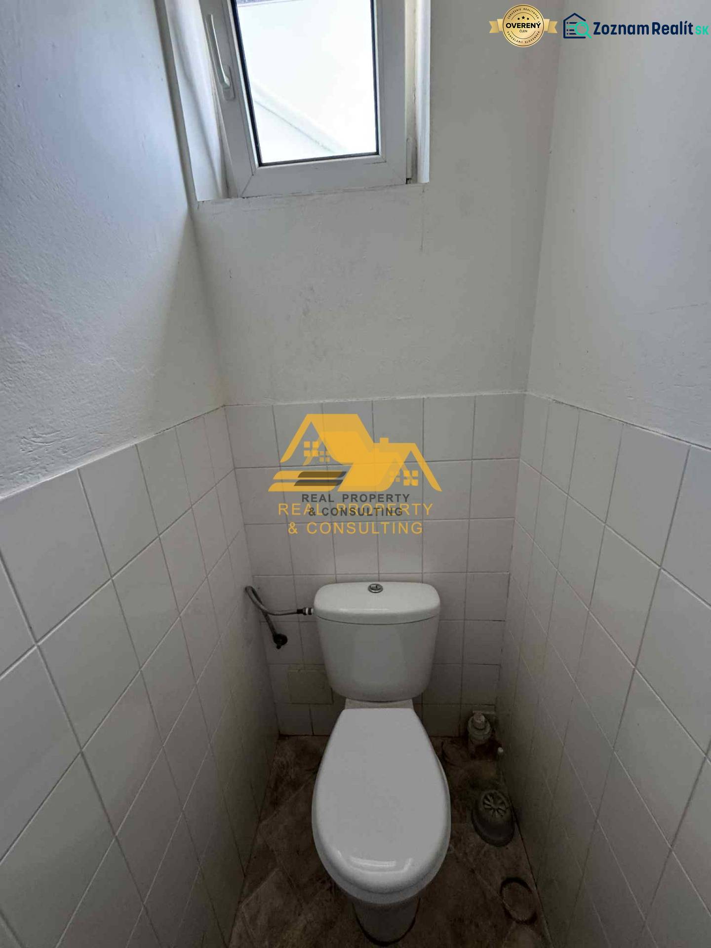 Kompakte Toilette mit Fenster, weiße Fliesen, Boden mit Holzdekor.