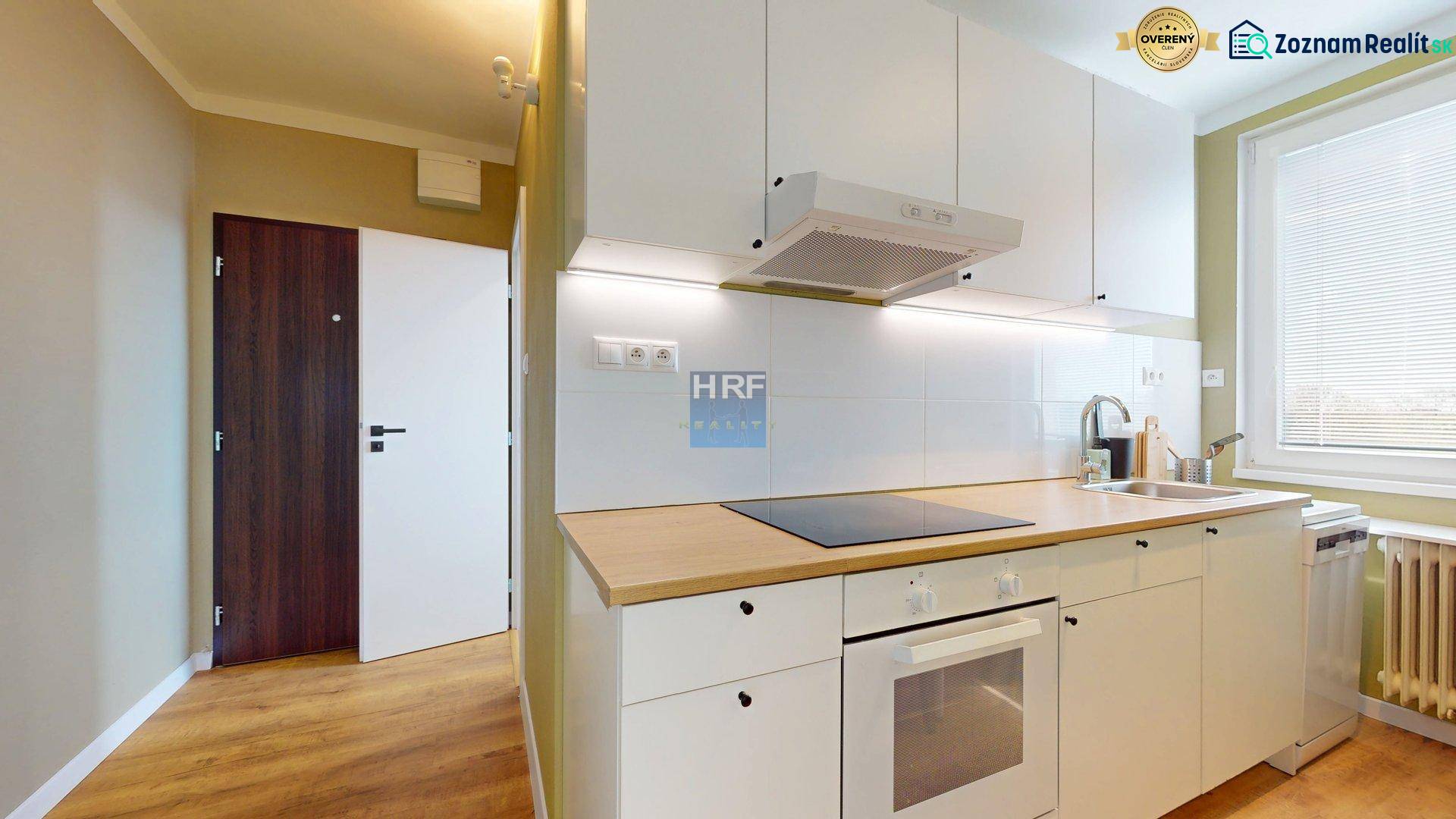 Küche in einer Einzimmerwohnung mit weißen Schränken, Boden mit Holzdekor, Kühlschrank, Kochfeld.