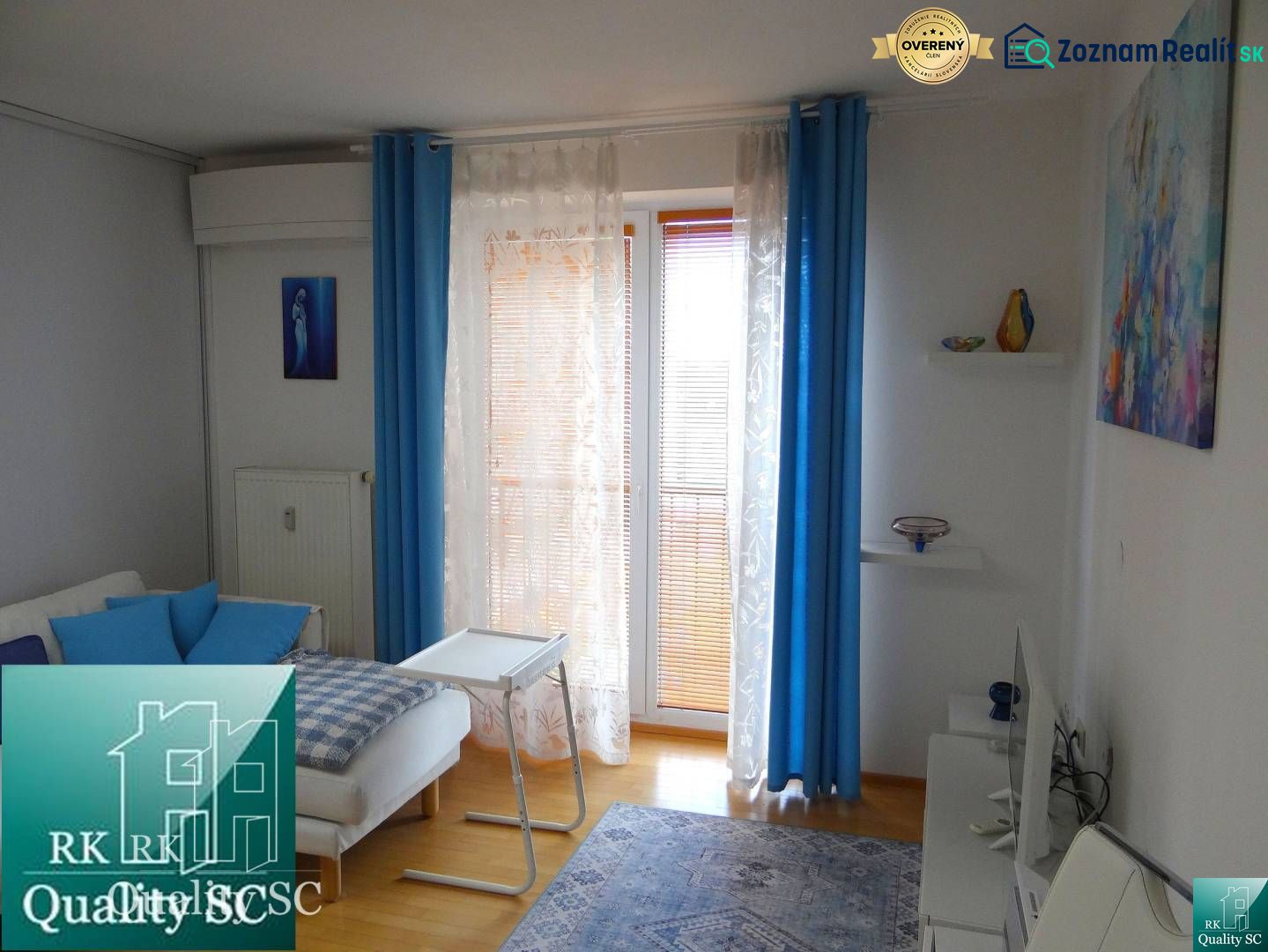 Ein Zimmer in einer Einzimmerwohnung mit weißen Wänden, einem Boden in Holzoptik und blauen Vorhängen.