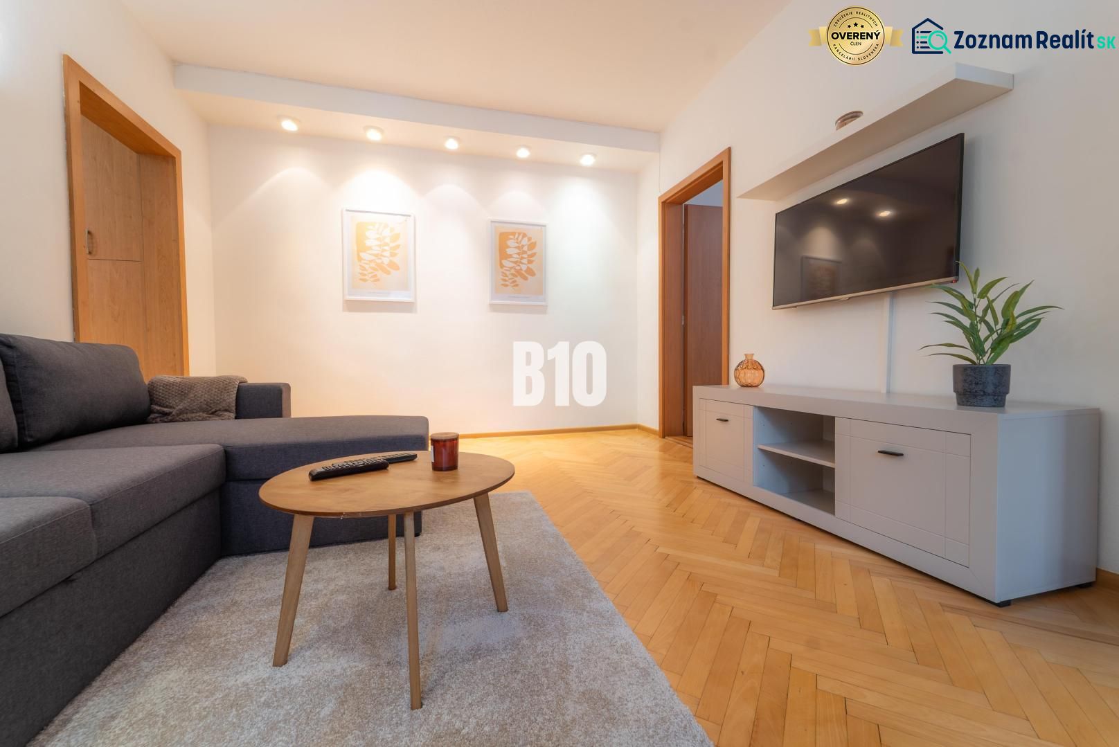 Wohnzimmer mit einem Fernseher an der Wand, einem grauen Sofa und einem Boden in Holzoptik.