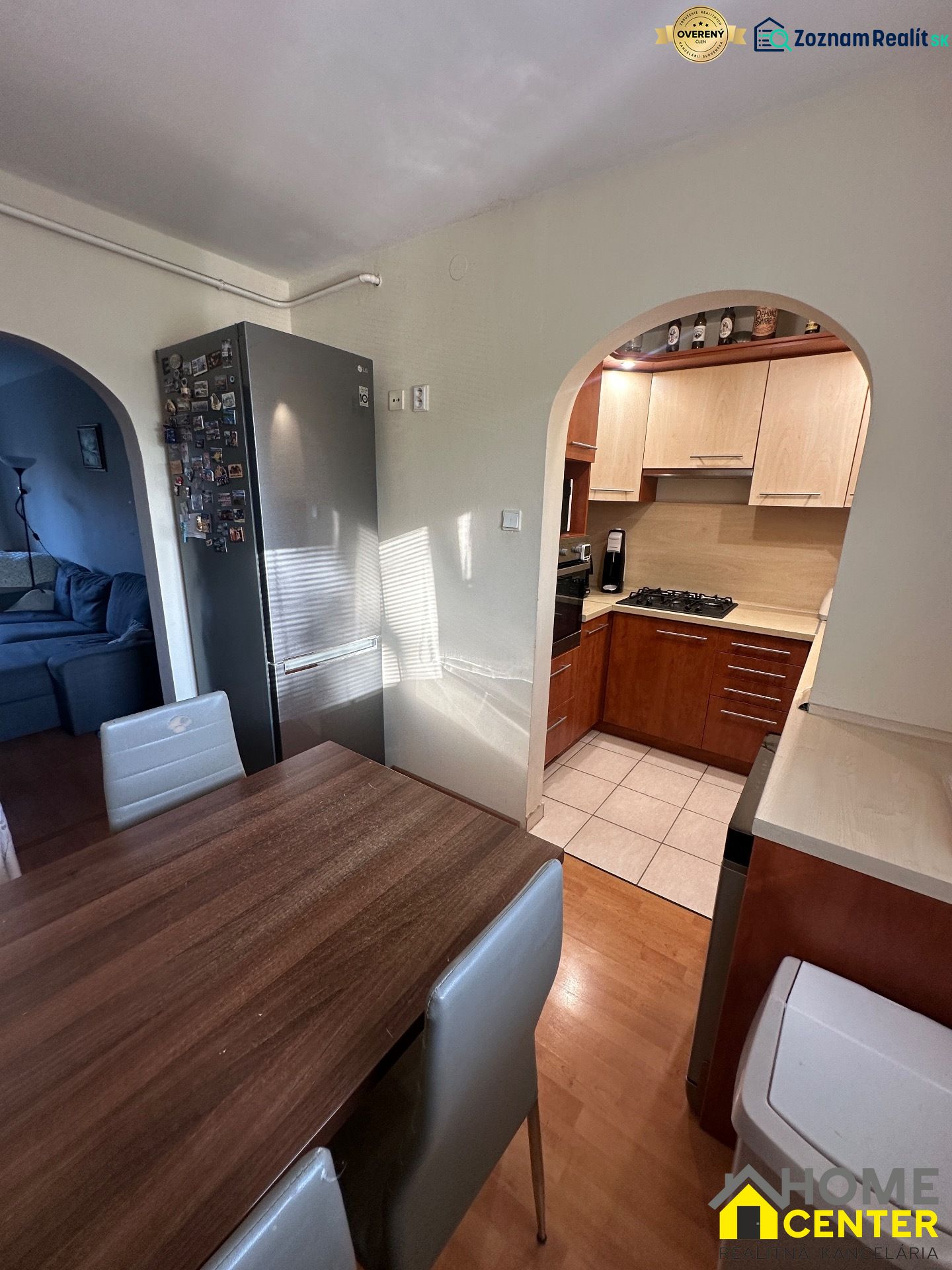 Küche mit Holzelementen, Esstisch, Kühlschrank und ein Bogen, der ins Wohnzimmer führt.