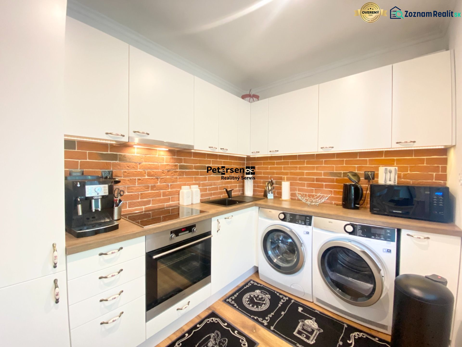 Küche in einer 2-Zimmer-Wohnung mit weißen Möbeln, Ziegelverkleidung und Geräten.