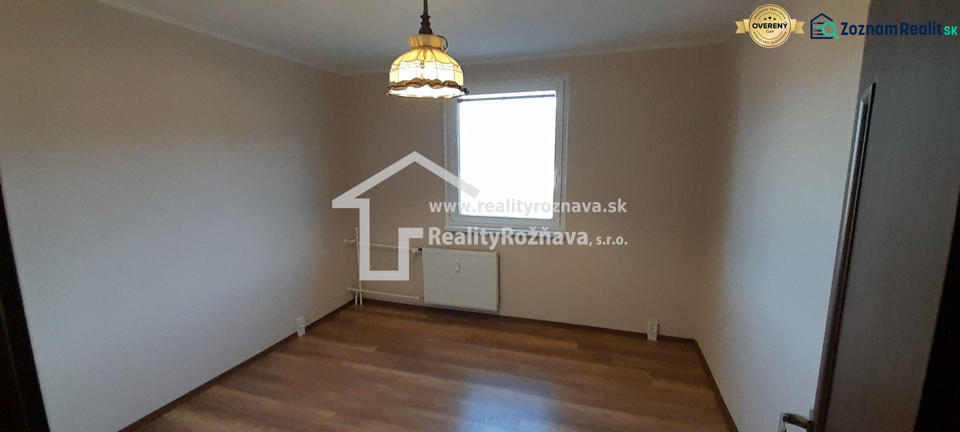 Ein Zimmer in einer 2-Zimmer-Wohnung mit einem Boden in Holzdekor und einer Lampe über dem Heizkörper.