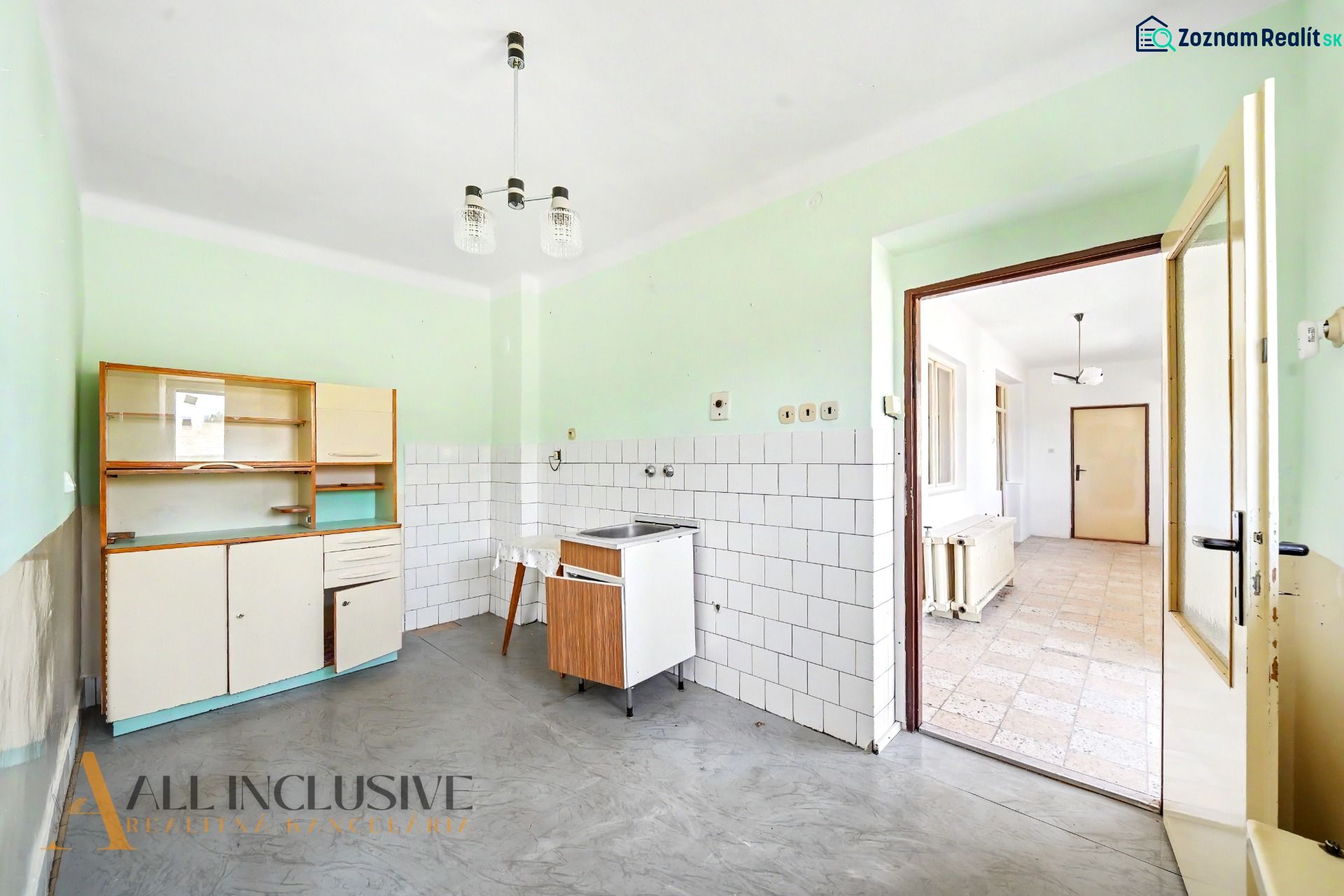Eine Küche in einem Einfamilienhaus mit einer einfachen Küchenzeile, weißen Fliesen und einem grauen Boden.