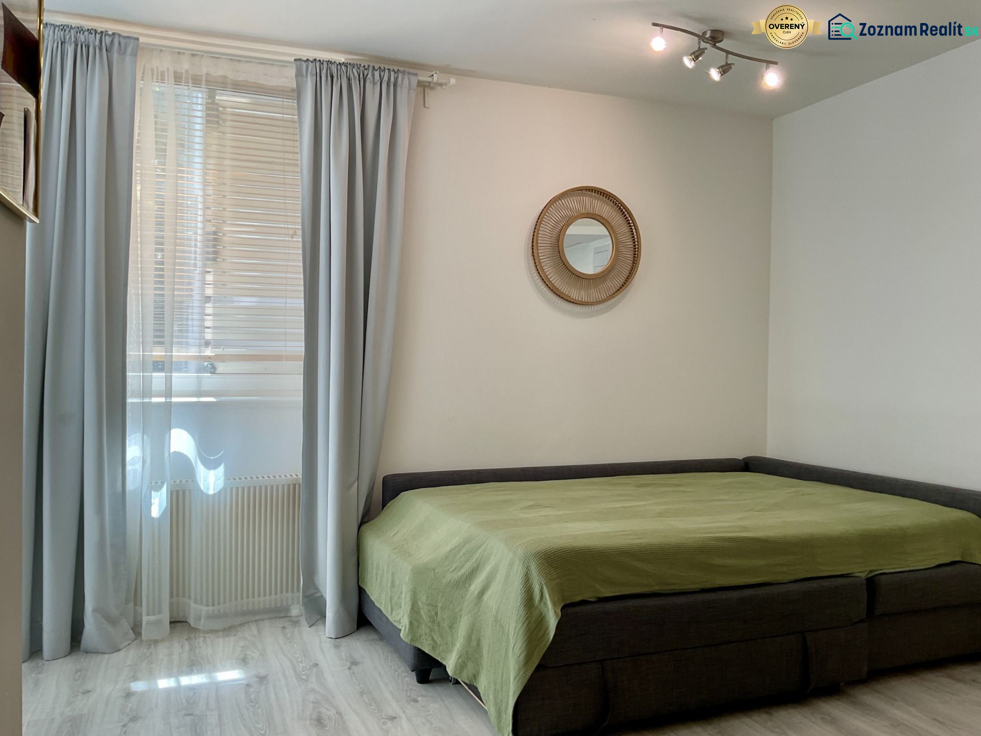 Ein Zimmer in einer Einzimmerwohnung mit einem Bett, Vorhängen und einem Boden in Holzoptik.