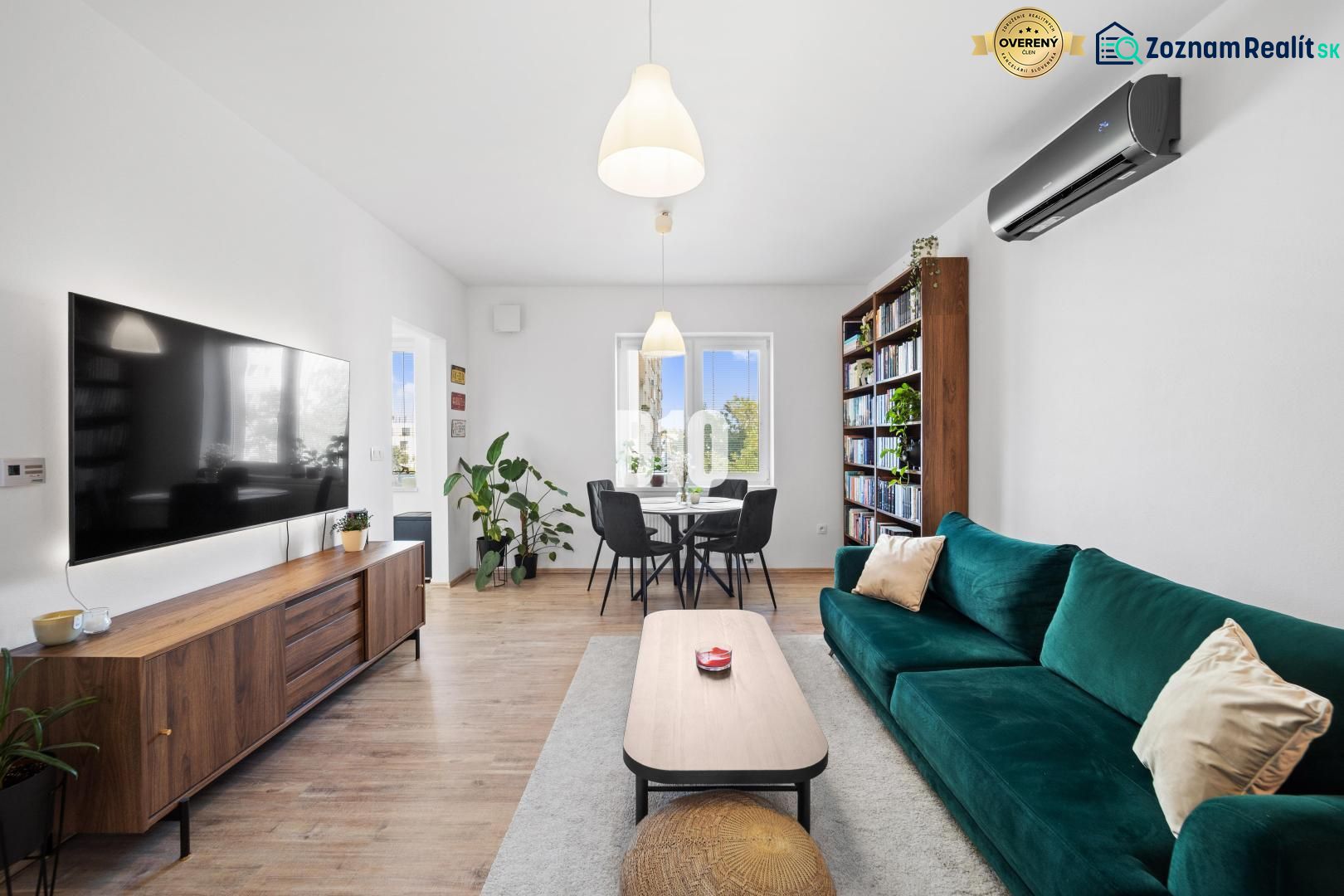 Wohnzimmer in einer Zweizimmerwohnung mit einem großen Fernseher, einem Bücherregal und einem Boden in Holzoptik.