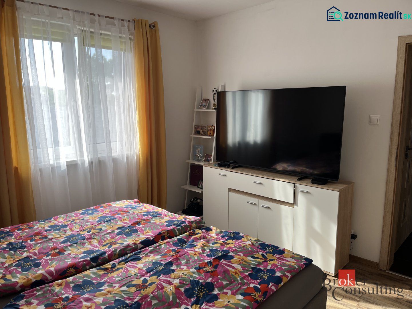 Schlafzimmer in einem Einfamilienhaus mit Fernseher, Blumenbettwäsche, gelbe Vorhänge, Fenster.