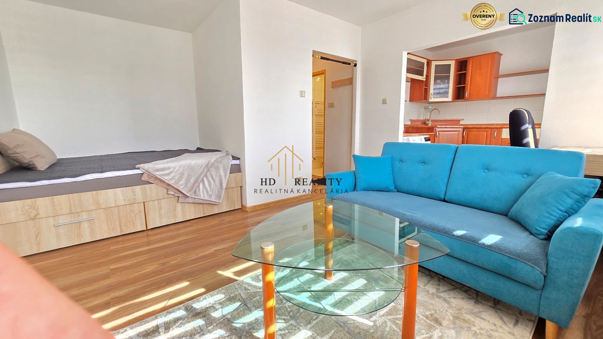 Ein Zimmer mit einem Holzboden, einem blauen Sofa und einer Kochnische.