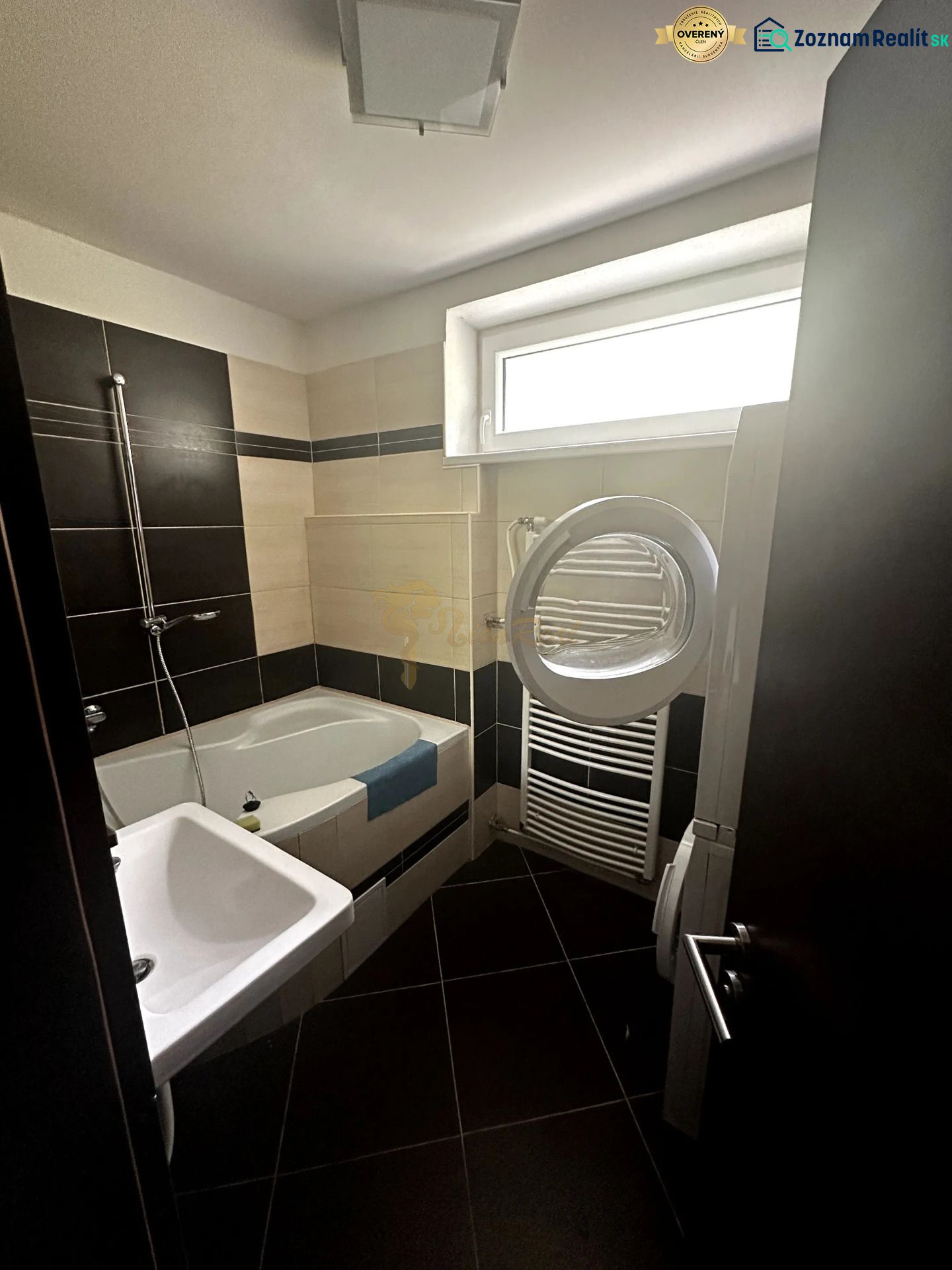 Badezimmer in einer 2-Zimmer-Wohnung mit Badewanne, Waschbecken und Waschmaschine; schwarze und beige Fliesen.
