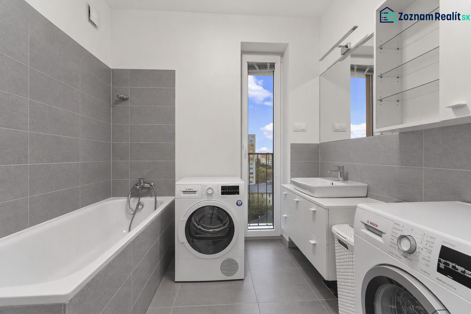 Badezimmer mit Badewanne, Waschmaschinen und Waschbecken in einer Zweizimmerwohnung. Graue Fliesen, großer Spiegel.