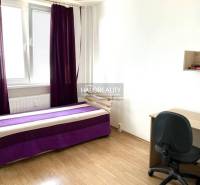 Zimmer: Bett mit lila Decke, Schreibtisch, schwarzer Stuhl, Fenster mit lila Vorhängen.