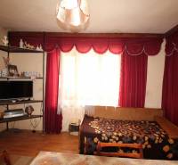 Wohnzimmer in einem Einfamilienhaus, mit roten Vorhängen, einem Bett und einem Regal mit Souvenirs.