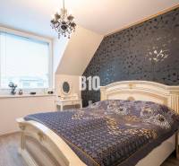 Ein Schlafzimmer in einem Einfamilienhaus mit dekorativer Tapete, elegantem Kronleuchter und Fenstern mit Jalousien.
