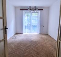 Ein leerer Raum mit Holzboden, einem großen Fenster und einem Kronleuchter in einer 2-Zimmer-Wohnung.