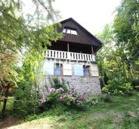 Eine Hütte in Holčíkovce, umgeben von Bäumen und Blumen, mit Steinfundamenten und einem weißen Balkon.