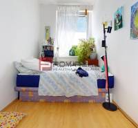 Ein Zimmer in einer 3-Zimmer-Wohnung mit weißen Wänden, einem Bett und einem Fenster mit Blumen.