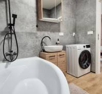 Ein Badezimmer mit grauen Fliesen, einer Badewanne, einer Waschmaschine und einem Waschbecken in einer Vierzimmerwohnung.