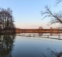 Ein See mit einem Steg, umgeben von kahlen Bäumen und im Hintergrund die warmen Farben des Sonnenuntergangs.