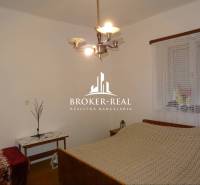 Schlafzimmer im Einfamilienhaus mit Bett, Tisch und Kronleuchter, an der Wand ist ein Fenster mit Gardine.