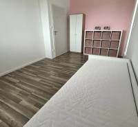 Ein helles Zimmer mit einer rosa Wand, einem Bett und einer Kommode, Boden mit Holzdekor.