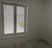 Ein Zimmer in einem Einfamilienhaus mit einem großen Fenster und Jalousien, weiße Wände, grauer Boden.