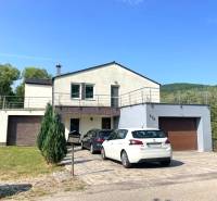 Ein Einfamilienhaus in Melčice-Lieskové mit einer Garage, zwei Autos und einem Garten auf einem Hanggrundstück.
