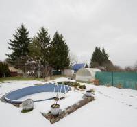 Ein verschneiter Garten bei einer Hütte in Handlová mit einem Pool, Gewächshäusern und Nadelbäumen.