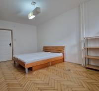 Schlafzimmer mit einem einfachen Bett, Regalen und einem Boden mit Holzdekor in einer 2-Zimmer-Wohnung.