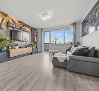 Wohnzimmer mit einem bequemen Sofa, einer Tapete mit Pflanzenmotiv und einem Boden in Holzoptik.