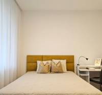 Schlafzimmer in einer Einzimmerwohnung mit hellem Bett, Vorhängen und einem weißen Tisch mit Lampe.