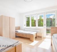 Ein heller Raum mit drei Betten, weißen Wänden, Schränken und großen Fenstern.