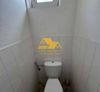 Kompakte Toilette mit Fenster, weiße Fliesen, Boden mit Holzdekor.