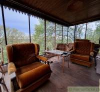 Eine Veranda mit Stühlen und einem Boden in Holzoptik, eine Hütte umgeben von Grün.