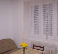 Ein Zimmer mit einem Sofa, einem Tisch, einem Stuhl und einem Vorhang, auf dem Tisch eine Vase mit Blumen.