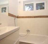 Badezimmer mit Badewanne und Waschbecken in einer Einzimmerwohnung, weiße Fliesen, Holzelemente.