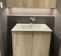 Kompaktes Badezimmer mit Schrank und Waschbecken in einer Einzimmerwohnung, helles Holz und dunkle Fliesen.