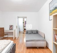 Helles Zimmer mit Bett, Tisch, Regal und Boden in Holzoptik.