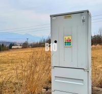 Ein elektrischer Schrank auf einer Wiese, im Hintergrund Hügel und ländliche Bebauung.