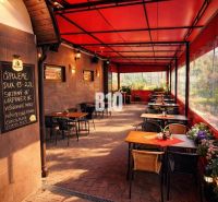Überdachte Terrasse des Restaurants im Gebäude der Stadt Čadca mit Ziegelpflaster, Blumen und Tischen.