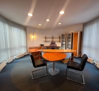 Ein Büro mit einem Tisch in Holzoptik, Regalen und beleuchteten vertikalen Jalousien.