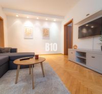 Wohnzimmer mit einem Fernseher an der Wand, einem grauen Sofa und einem Boden in Holzoptik.